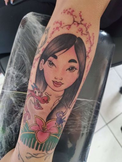 Color forearm tattoo of an animated Mulan character with a small red dragon and cherry blossoms.
