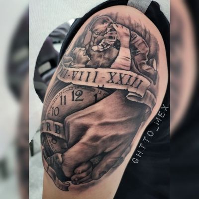 Black and grey upper arm tattoo of a parent holding a newborn above a Roman numeral date banner and pocket watch
