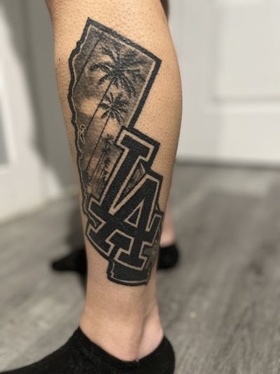 Black and grey calf tattoo of a California state outline filled with palm trees and clouds, behind a large interlocking LA.