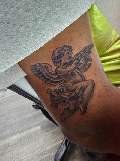Black and grey cherub with feathered wings aiming a rifle, tattooed on an upper arm
