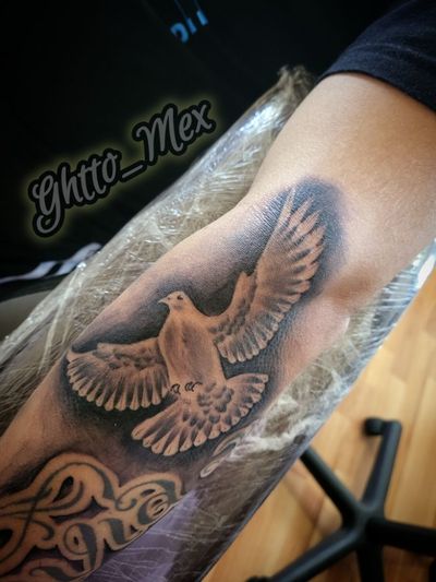 Black and grey dove with wings spread on a forearm, soft smoke shading behind it and script lettering below