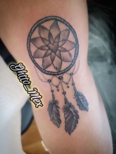 Black and grey dreamcatcher tattoo on an inner arm, layered petal web with a heart center and three hanging feathers