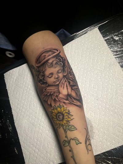 Black and grey praying cherub with a halo and wings above a color sunflower on the inner forearm, photographed fresh.