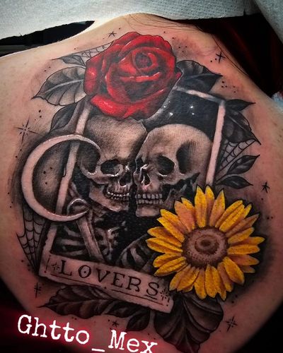 Upper back tattoo of two skeletons embracing inside a tarot card labeled Lovers, with a red rose and sunflower