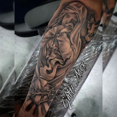 Black and grey forearm tattoo of the Virgin Mary praying with a rosary above a crown of thorns